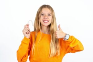 Teen girl holding clear aligner, making thumbs-up gesture